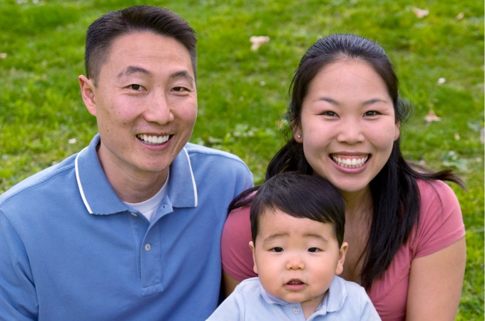 A family sits on the grass for a photo. You see two adults smiling at the camera with a young child seated in front of them