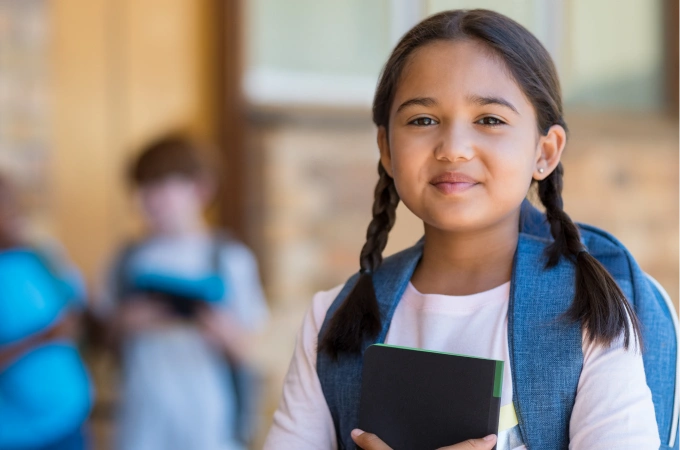 A young student stands outside a school building holding a notebook. She has braids, a backpack, and a confident, relaxed smile